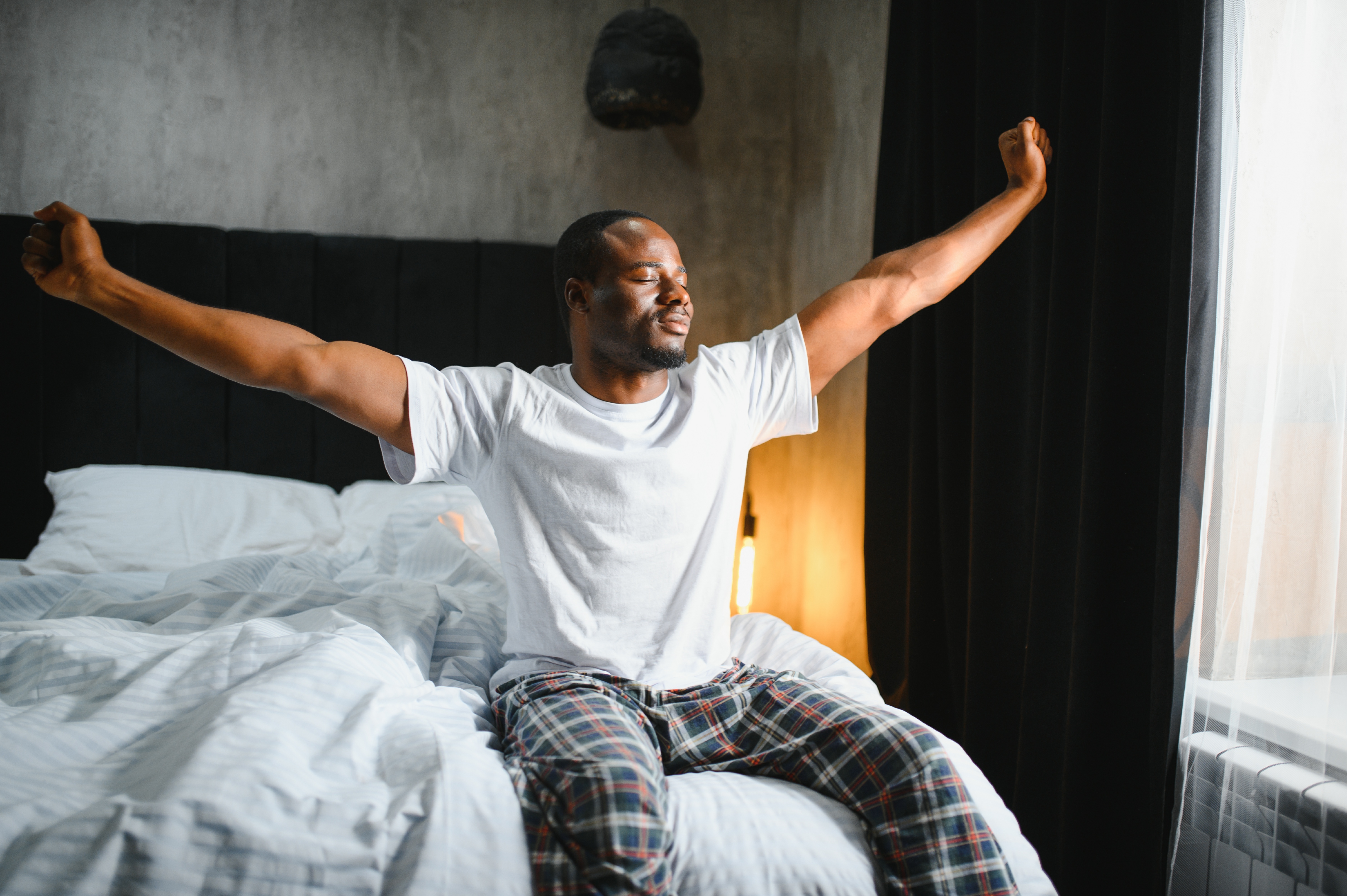Good Morning. Happy Black Man Waking Up And Stretching Arms With Smile, Relaxed After Great Rest, Sitting In Bed In Modern Bedroom Interior. Start Of New Day, Rest And Wellness Concept