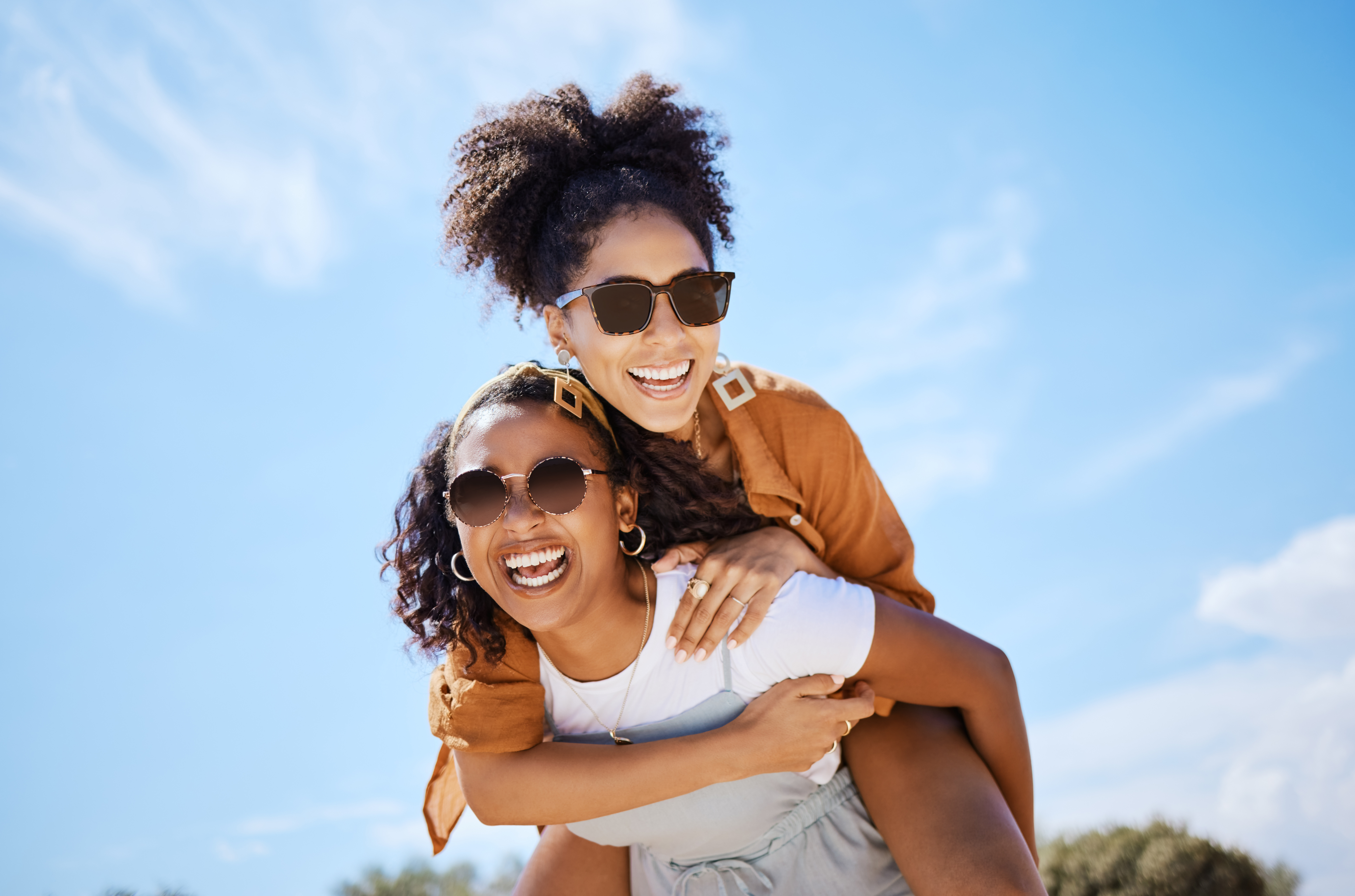 Summer, friends and freedom with women and piggy back against the blue sky for hug, lifestyle and happy on Puerto rico holiday. Youth, crazy and friend with girl on vacation with glasses and a smile