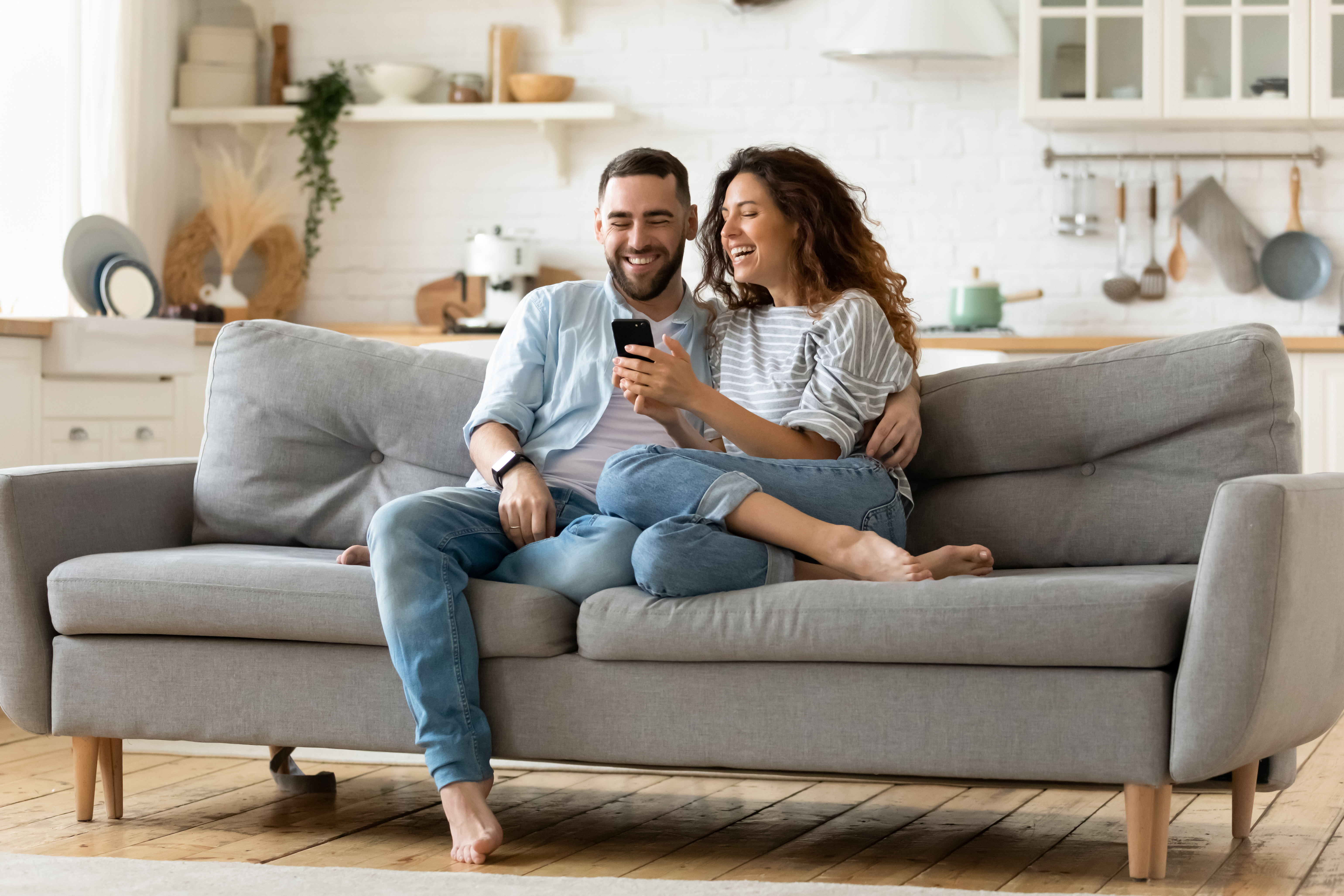 Happy young woman and man hugging, using smartphone together Happy young woman and man hugging, using smartphone together, sitting on cozy couch at home, smiling overjoyed wife and husband looking at phone screen, sitting on sofa in modern living room