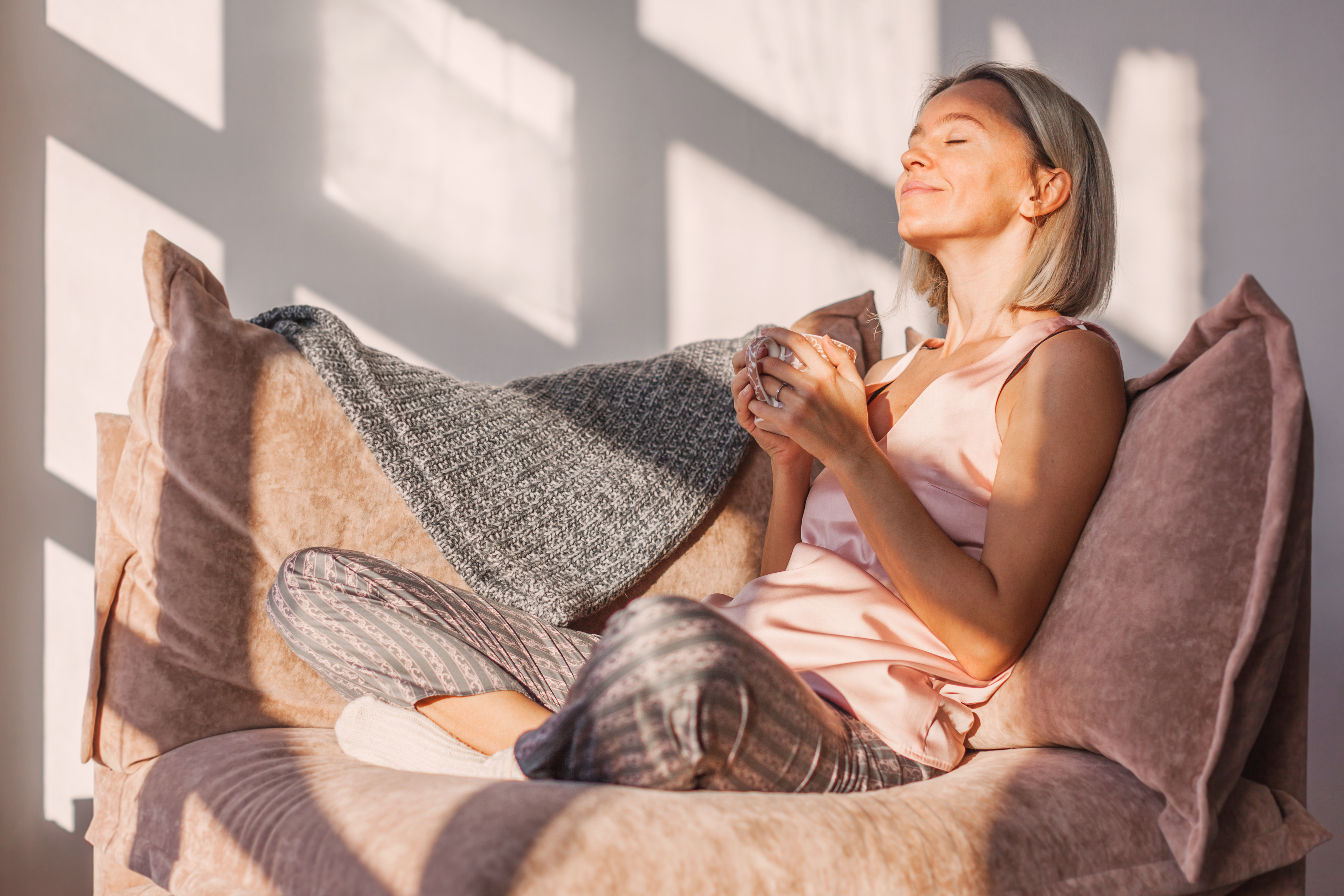 Happy dreamy middle aged woman sitting on sofa in living room wi Adult woman sitting on the sofa at home and having a cup of coffee, relaxation concept